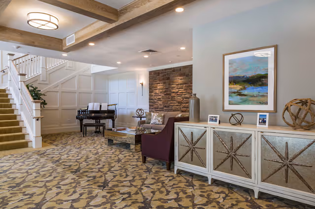 A decorated communal lounge with patterned carpet, seating, a grand piano, staircase, and a decorative sideboard under framed art.