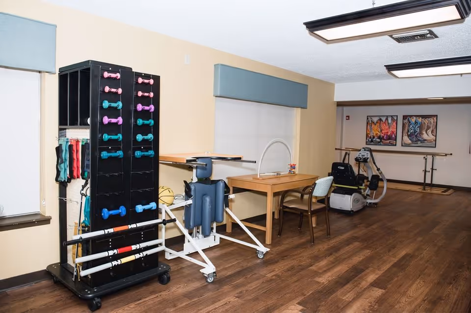 A rehabilitation room with exercise equipment including a rack of colorful hand weights, a therapy table with a chair, a recumbent exercise bike, and parallel bars along the back wall. The room has wooden flooring, beige walls, and framed pictures of cowboy boots on the far wall.