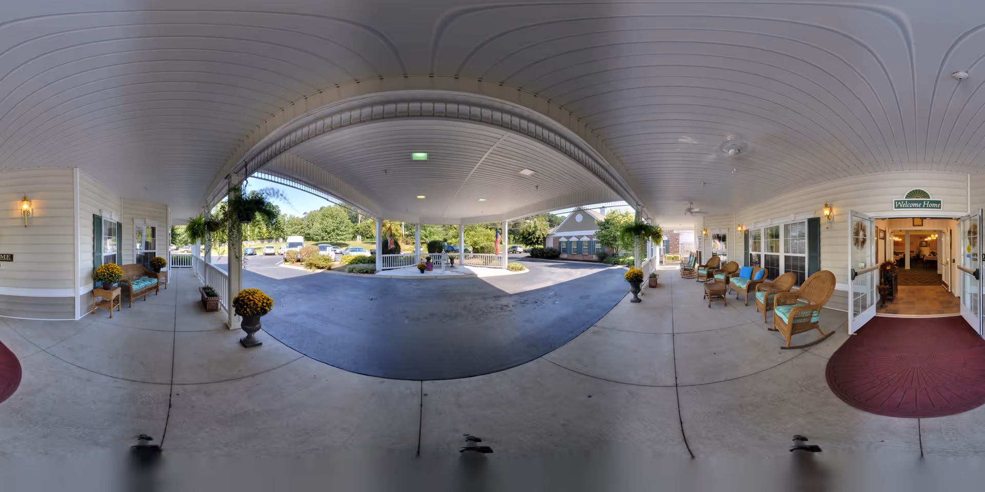 Covered entrance area of Charter Senior Living of Jackson with a circular driveway. There are wicker chairs with cushions lined up along the right side under the covered porch, potted plants with yellow flowers, and a 'Welcome Home' sign above the open double doors leading inside. The area is well-lit with wall-mounted lantern-style lights and ceiling lights. Outside the covered area, there are trees, shrubs, and parked vehicles visible.