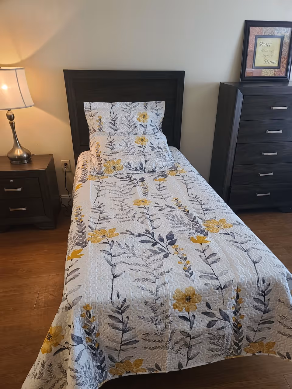 A single bed with a yellow-and-gray floral quilt, a nightstand and lamp to the left and a dresser with framed art to the right.