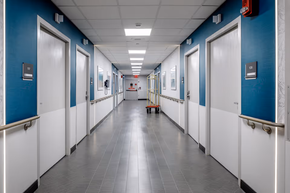 A clean, well-lit hallway in a senior living facility with white doors on both sides, blue and white walls, handrails along the walls, ceiling lights, a fire alarm, and a luggage cart near the end of the corridor.