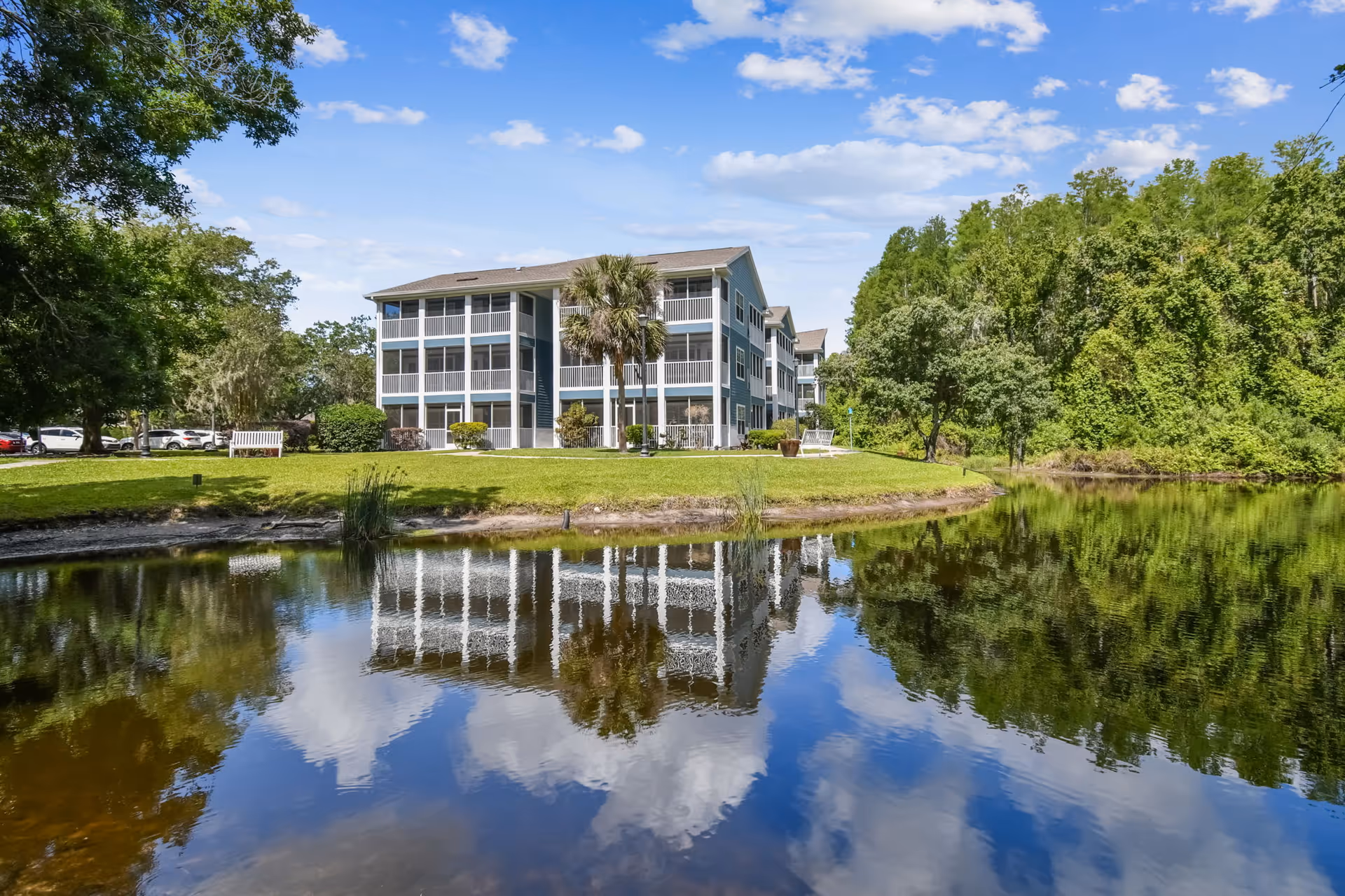 A multi-story residential building with screened balconies is situated beside a calm pond reflecting the building, blue sky, and clouds. The area around the building is landscaped with grass, trees, and shrubs, and there is a bench near the water's edge.