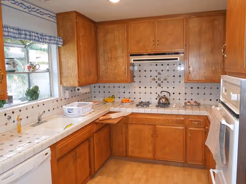 A kitchen with wooden cabinets, white tiled countertops and backsplash with black accents, a gas stove with a kettle, a white dishwasher, a white oven, a window with a blue and white checkered valance, and various kitchen items including a dish rack, bananas, and oranges.