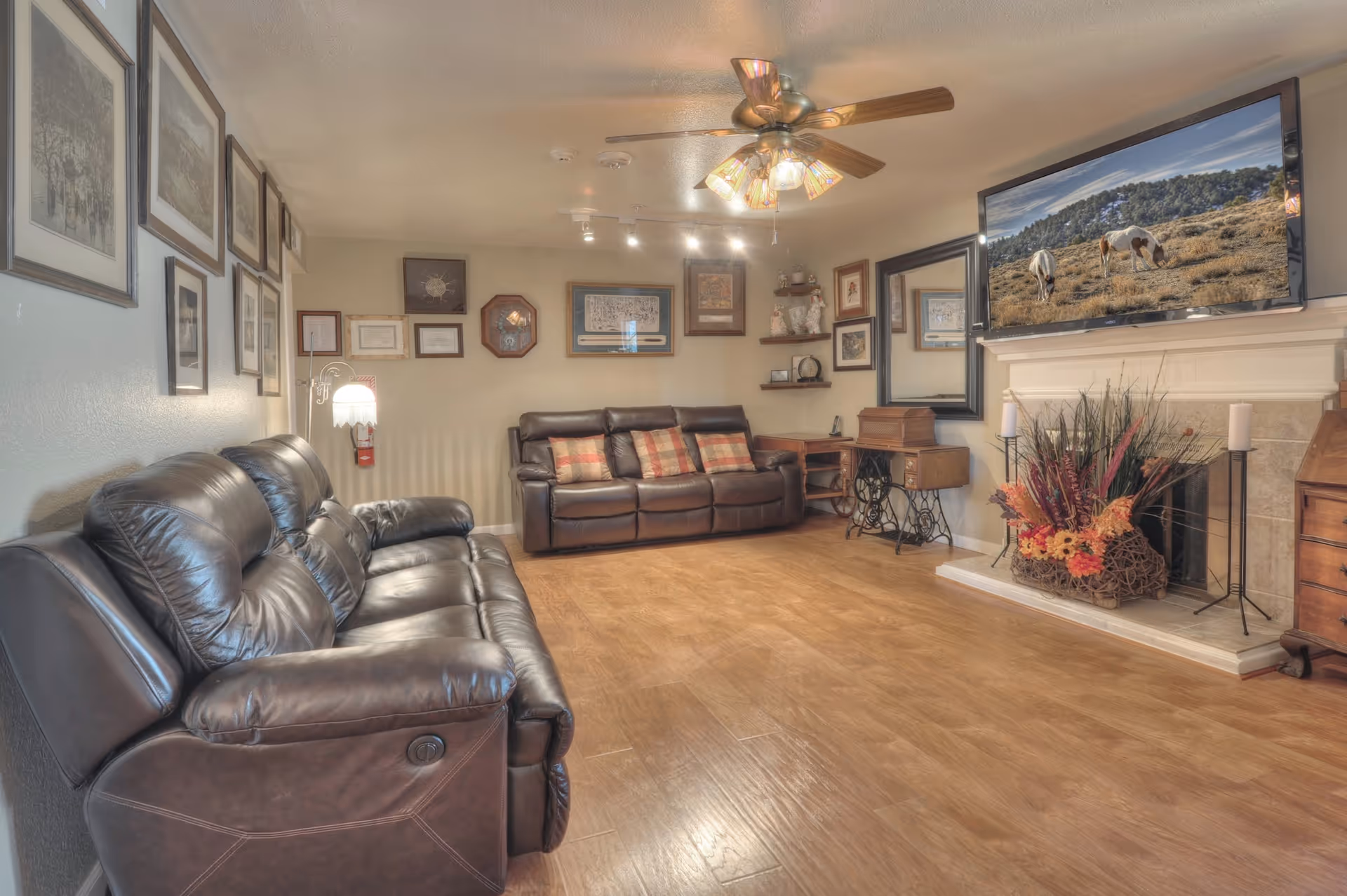 A cozy living room with two dark brown leather sofas, a ceiling fan with lights, framed pictures and certificates on the walls, a large flat-screen TV mounted above a white fireplace decorated with a floral arrangement, and wooden flooring.