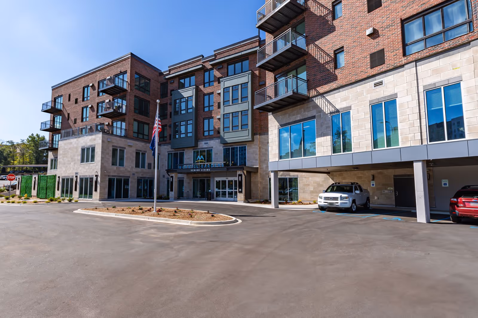 Exterior view of Merrill Gardens at Greenville, a multi-story senior living facility with brick and stone facade, balconies, large windows, an American flag on a flagpole, and a parking area with cars.