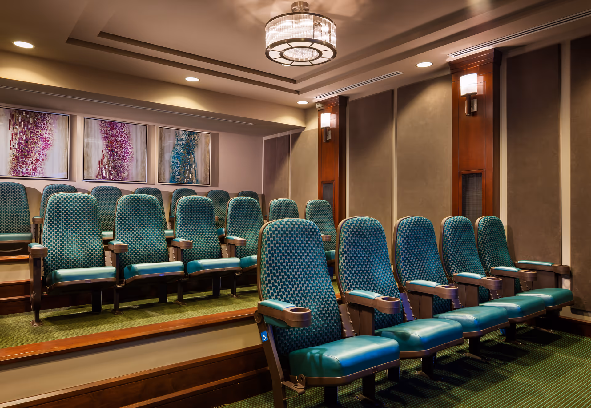 A small theater room with two rows of teal upholstered seats with wooden armrests, green carpeted floor, beige walls with wooden paneling, three abstract paintings on the back wall, and a modern ceiling light fixture.