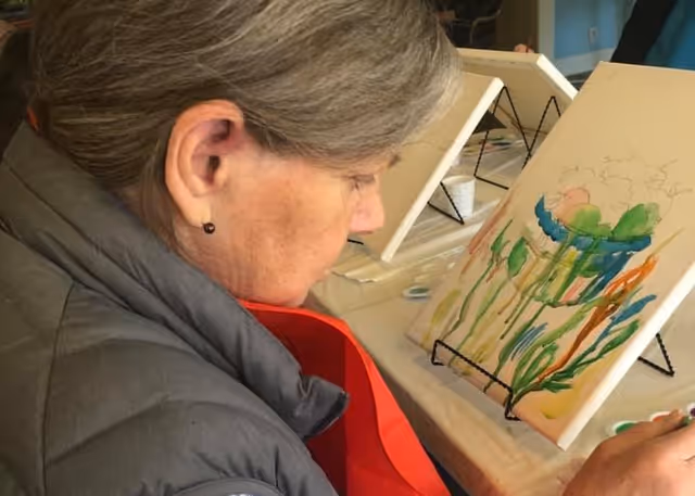 An elderly woman with gray hair wearing a gray jacket and red apron is painting a colorful floral design on a canvas set on a table easel indoors.