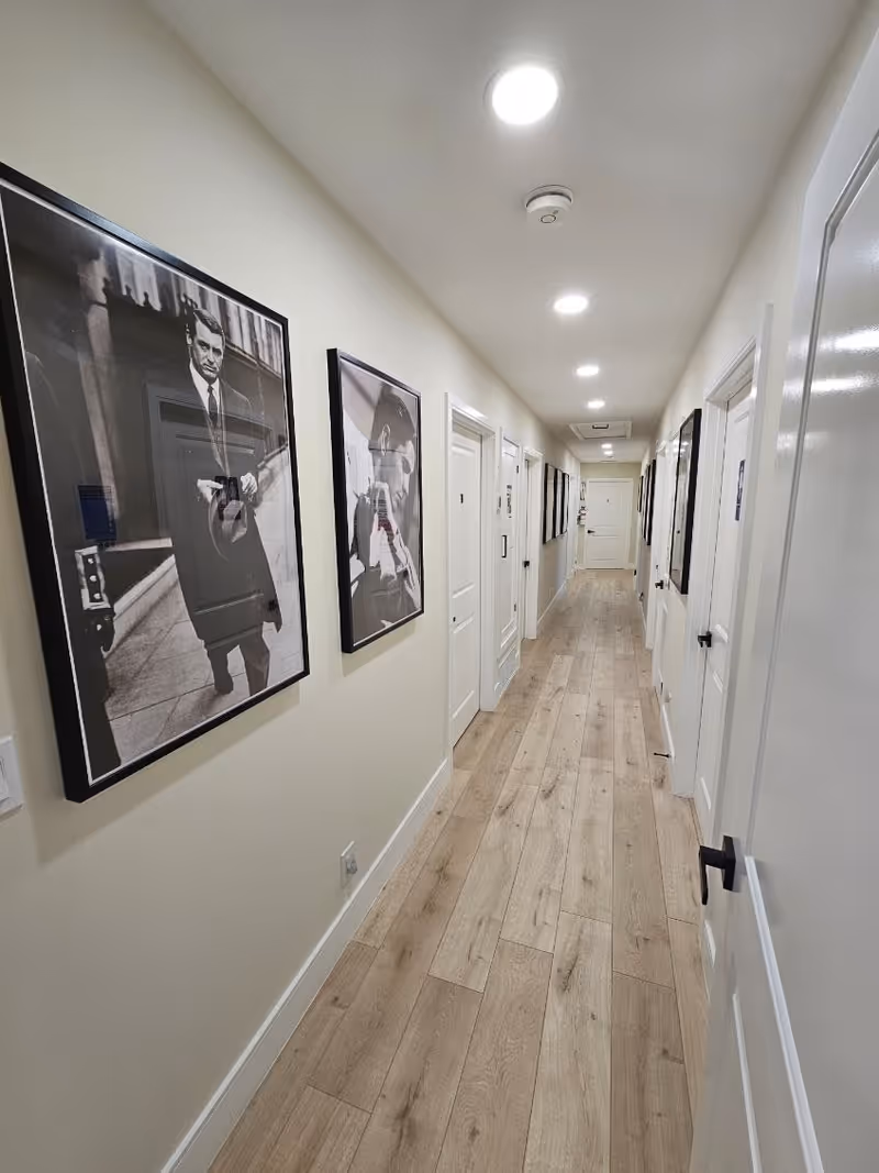 A long, narrow hallway with light wood flooring and cream-colored walls. Several white doors line both sides of the hallway, each with black handles. The ceiling has recessed lighting and a smoke detector. Black and white framed photographs hang on the left wall.
