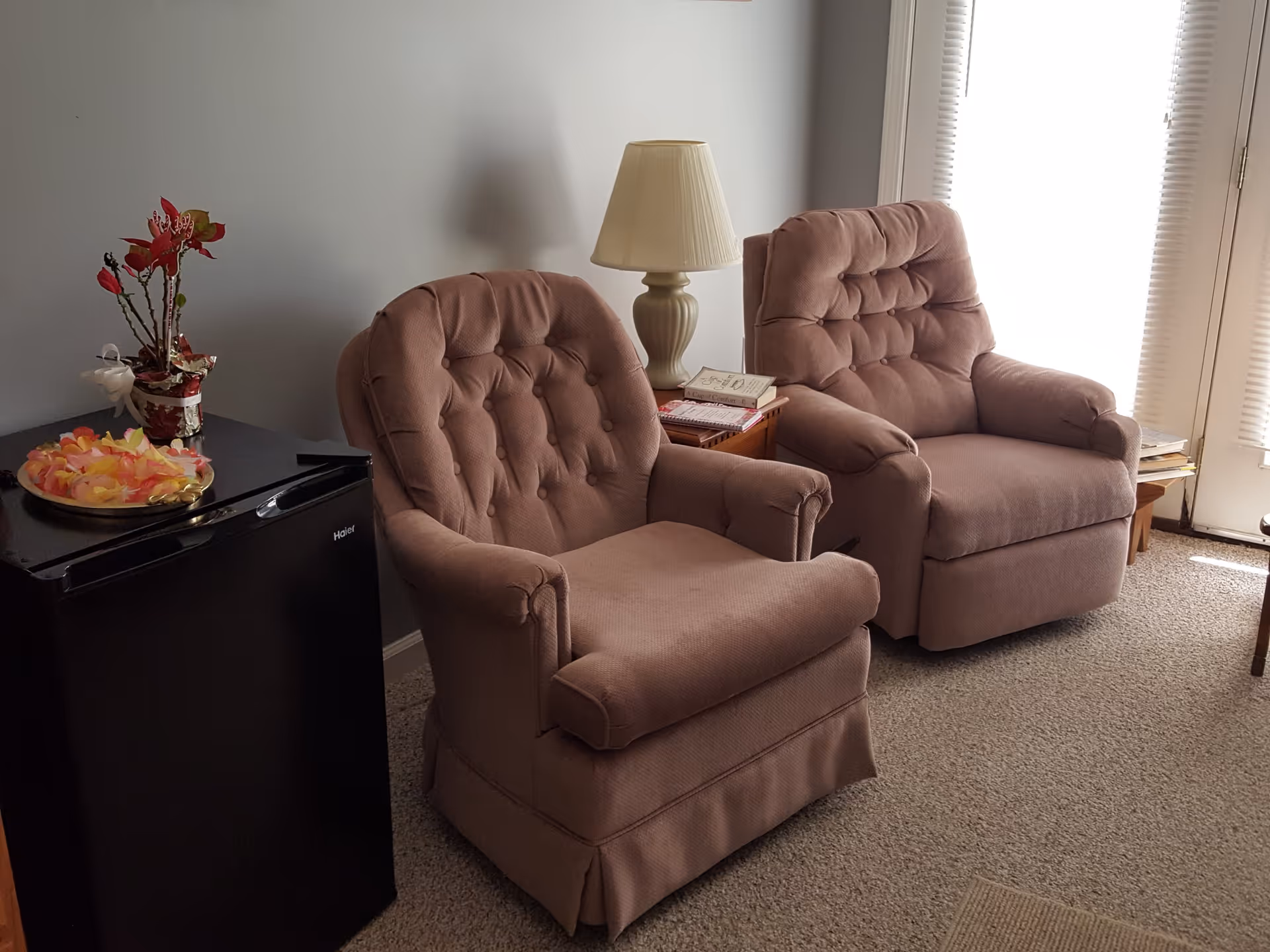 Two cushioned armchairs in a living area next to a small black refrigerator with a decorative flower arrangement and a plate of flower petals on top. A table lamp and some books are on a small wooden side table between the chairs. Light filters in through a window with blinds in the background.