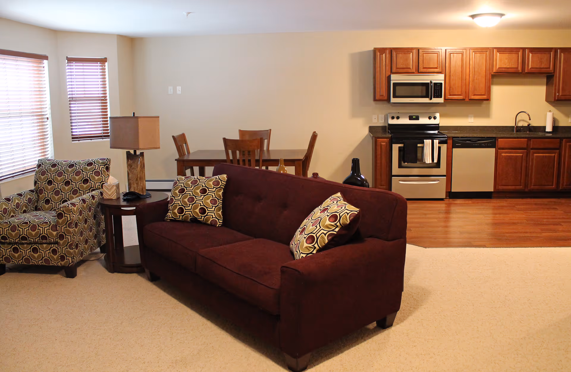 Interior view of a senior living facility showing a living room area with a maroon sofa and patterned armchair, a wooden side table with a lamp, a dining table with chairs in the background, and a kitchen area with wooden cabinets, stainless steel appliances including a stove, microwave, and dishwasher.