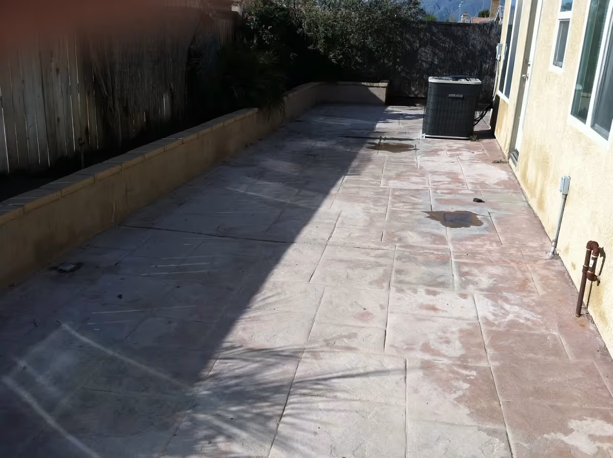 Outdoor patio area with tiled flooring, a raised planter bed along the left side with some plants, a wooden fence, and an air conditioning unit near the building wall on the right side.