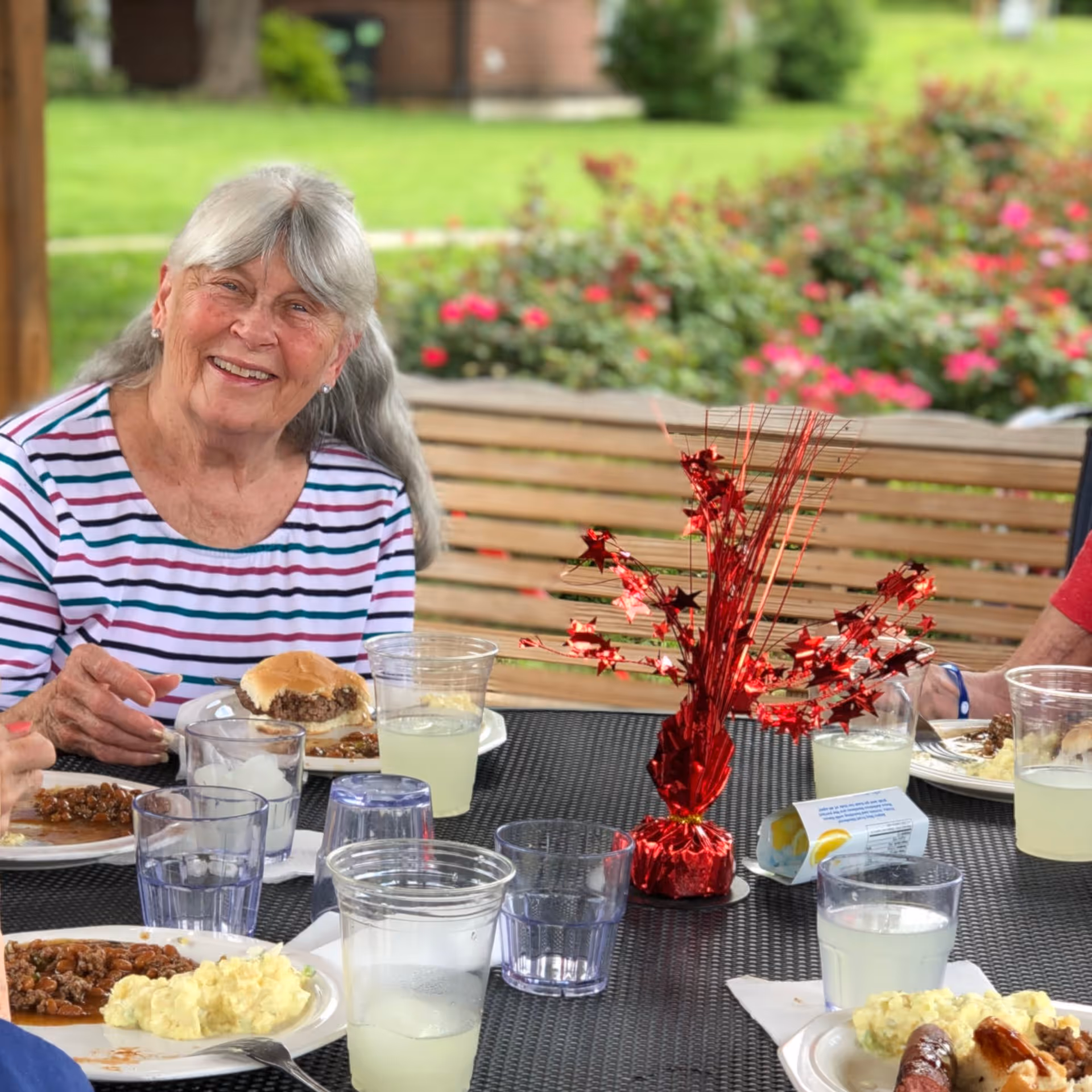 An elderly woman with gray hair wearing a striped shirt is smiling while sitting at a table outdoors. The table has plates of food including mashed potatoes, baked beans, and a hamburger, along with several clear plastic cups filled with lemonade or water. A red decorative centerpiece is on the table, and there are green grass and flowering bushes in the background.