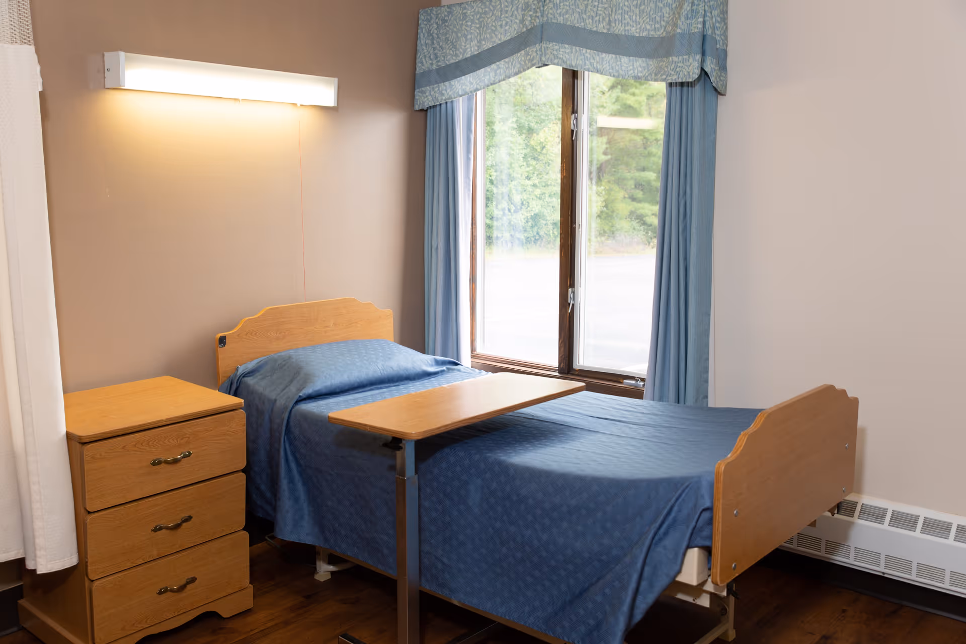 A small bedroom with a single hospital-style bed covered in a blue bedspread, a wooden bedside table with three drawers, a wooden overbed table, and a large window with blue curtains and a valance, showing greenery outside.