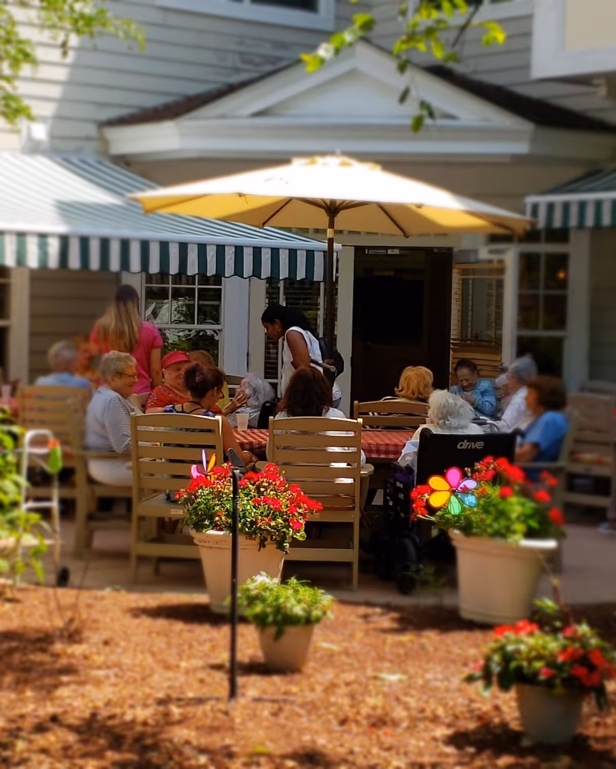 A group of elderly people sitting around a table with a red and white checkered tablecloth under a large yellow patio umbrella outside a building. There are several potted plants with red flowers in the foreground, and a caregiver or staff member is interacting with the group.