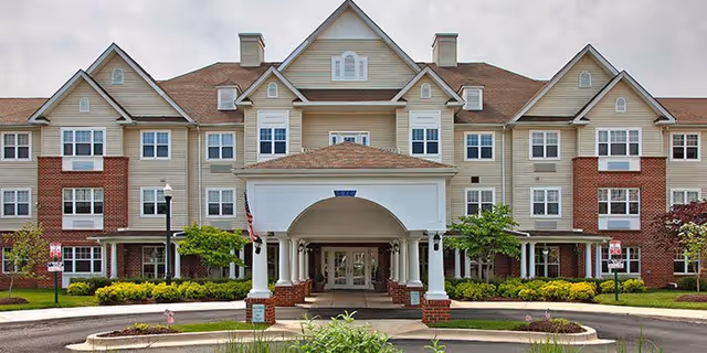 Front entrance of a multi-story senior living building with a covered porte-cochere, landscaped circular drive, and an American flag.