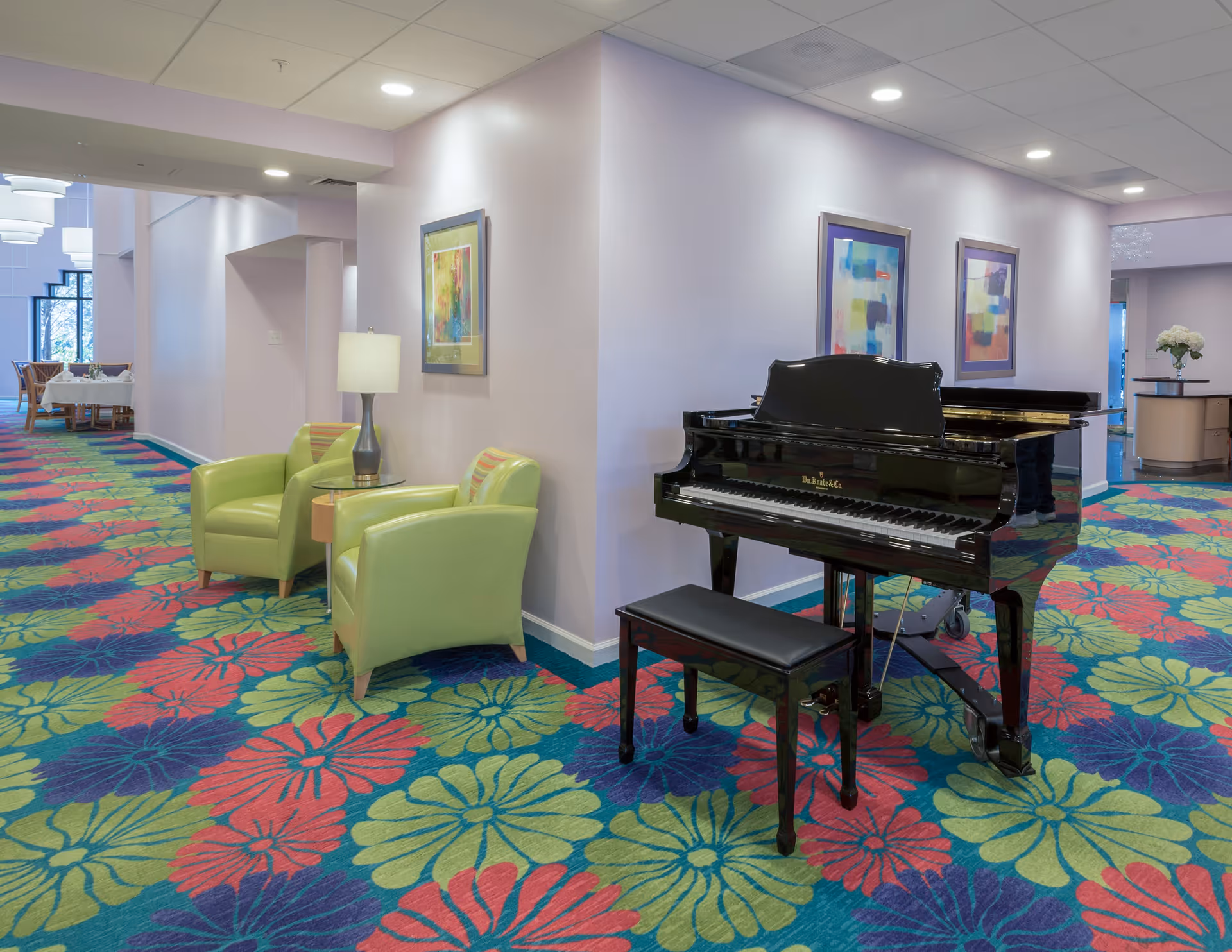 Interior common area of a senior living facility featuring a black grand piano with a bench, two green armchairs with a small round table and lamp between them, colorful floral patterned carpet, and abstract artwork on the walls.