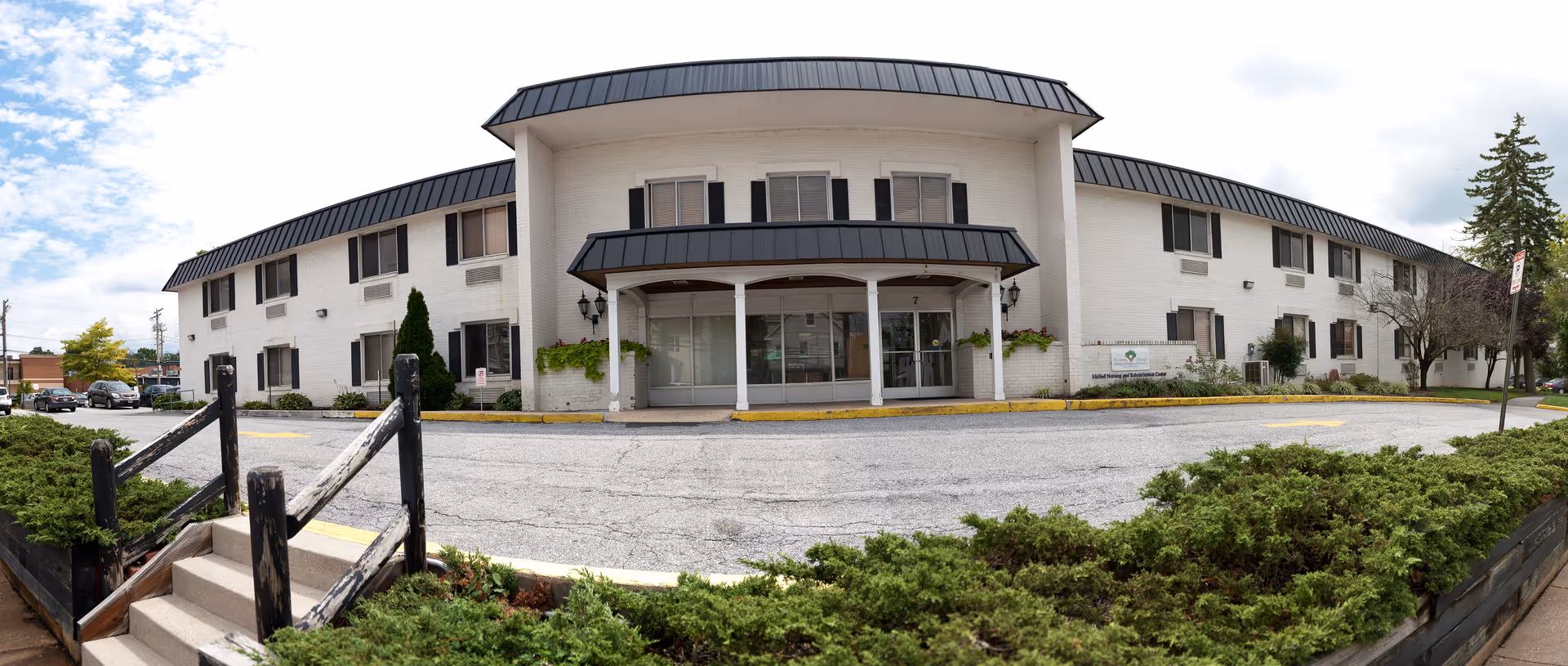 Front exterior view of a two-story white brick building with black trim and a covered entrance supported by white columns. There are shrubs and greenery in front of the building, a small staircase with a wooden railing leading up to the entrance area, and a parking lot in front. The sky is partly cloudy.