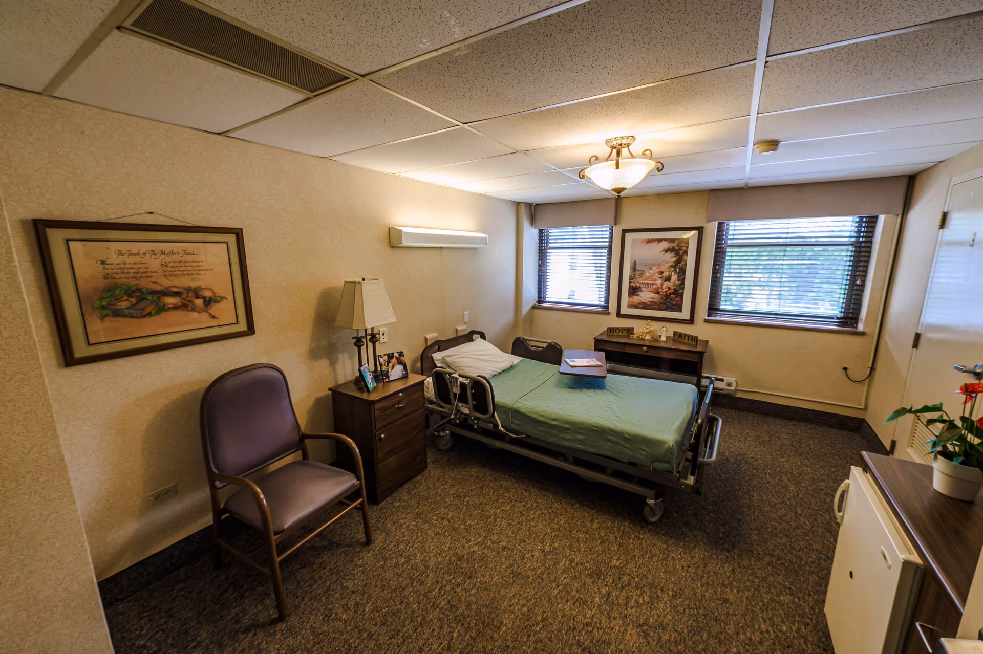 A small, cozy room with a hospital-style bed covered with a green sheet, a wooden nightstand with a lamp and framed photos, a purple armchair, two windows with blinds, a framed picture on the wall, and a small cabinet with a potted plant and a mini fridge.