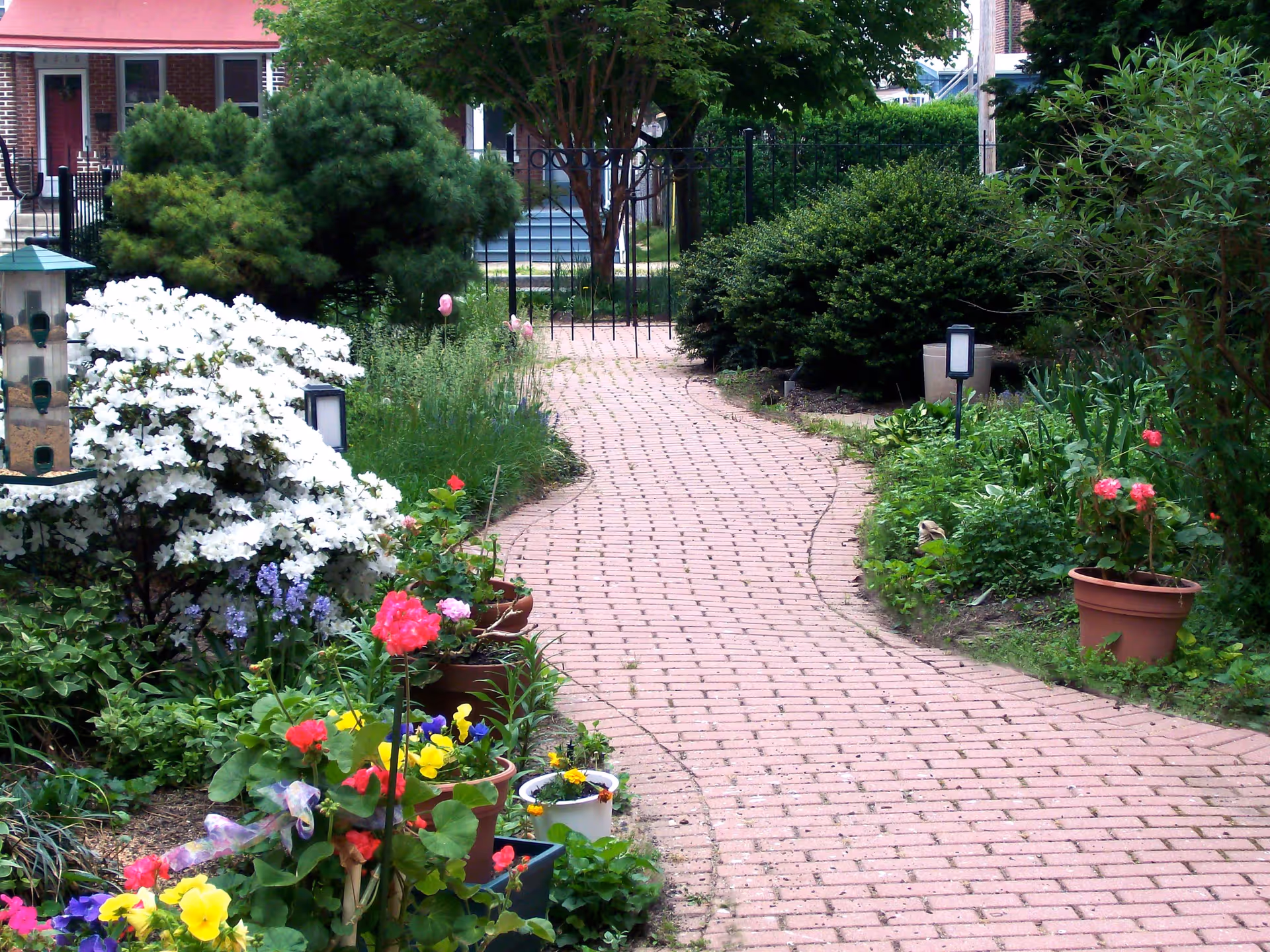 A winding brick pathway surrounded by lush greenery and colorful flowers in pots and garden beds, leading to a black metal gate with a red awning and building visible in the background.