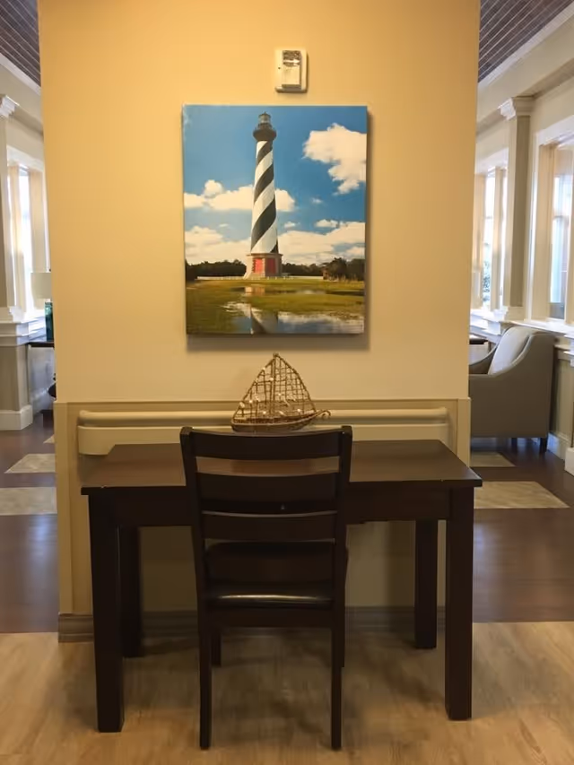 A small dark wooden desk with a matching chair is placed against a beige wall. On the desk is a decorative model of a sailboat. Above the desk hangs a large framed picture of a black and white striped lighthouse with a blue sky and clouds in the background. The surrounding area shows part of a well-lit interior with windows and seating areas.