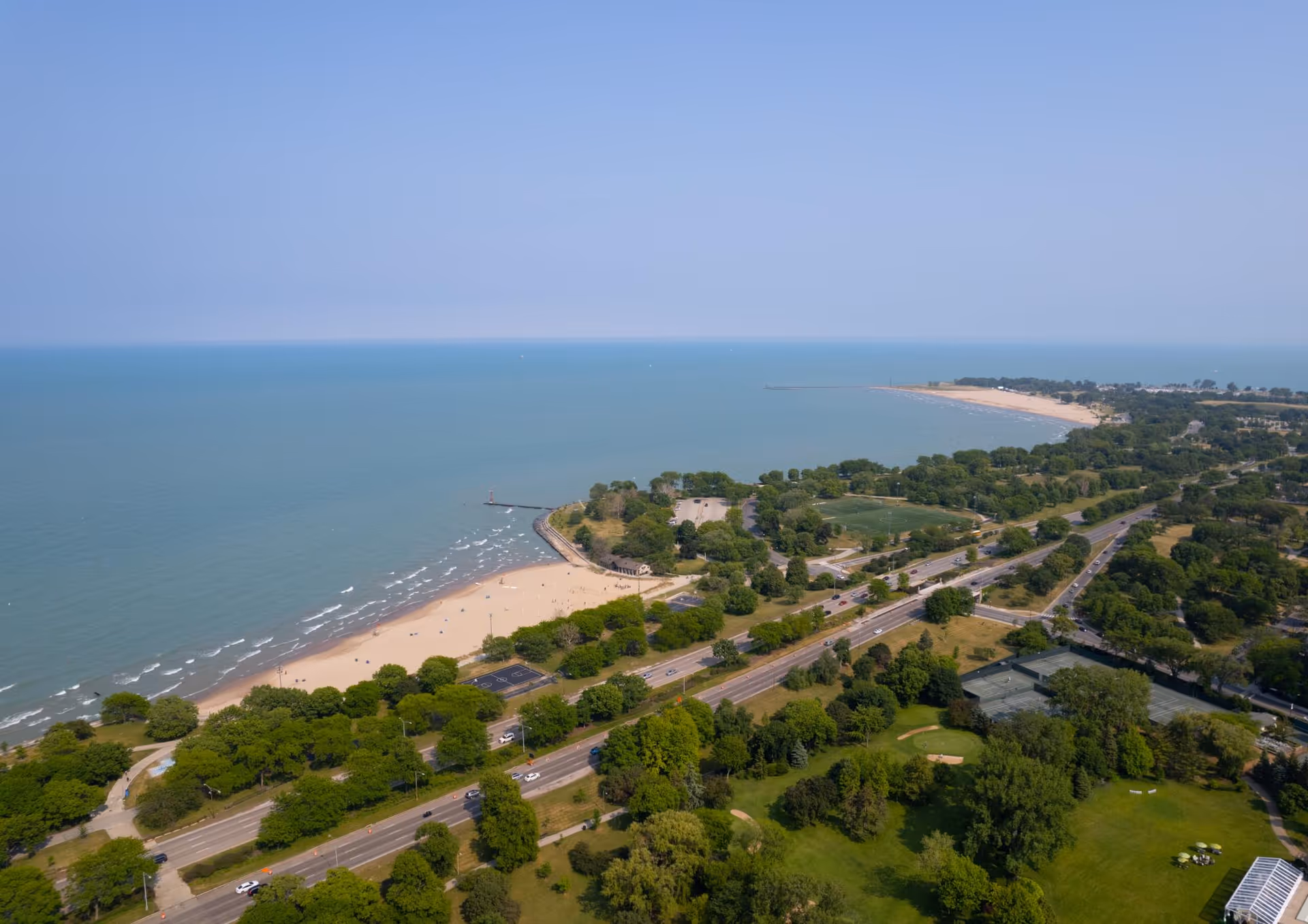 Aerial view of a coastal area showing a sandy beach, a large body of water, green trees, roads with cars, and open grassy spaces under a clear blue sky.