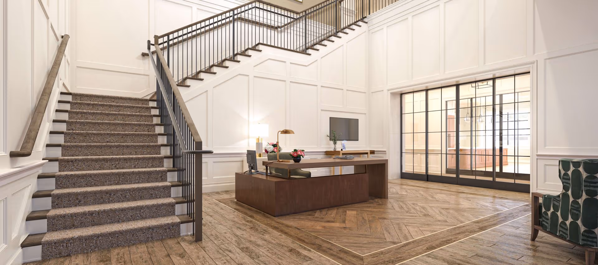 A spacious and elegant interior lobby area featuring a carpeted staircase with wooden handrails and black metal balusters. There is a modern wooden reception desk with a computer, desk lamp, and flower arrangements. The walls are white with decorative paneling, and the floor has a wood pattern. Large glass doors with black frames lead to another room, and a green patterned armchair is partially visible on the right.