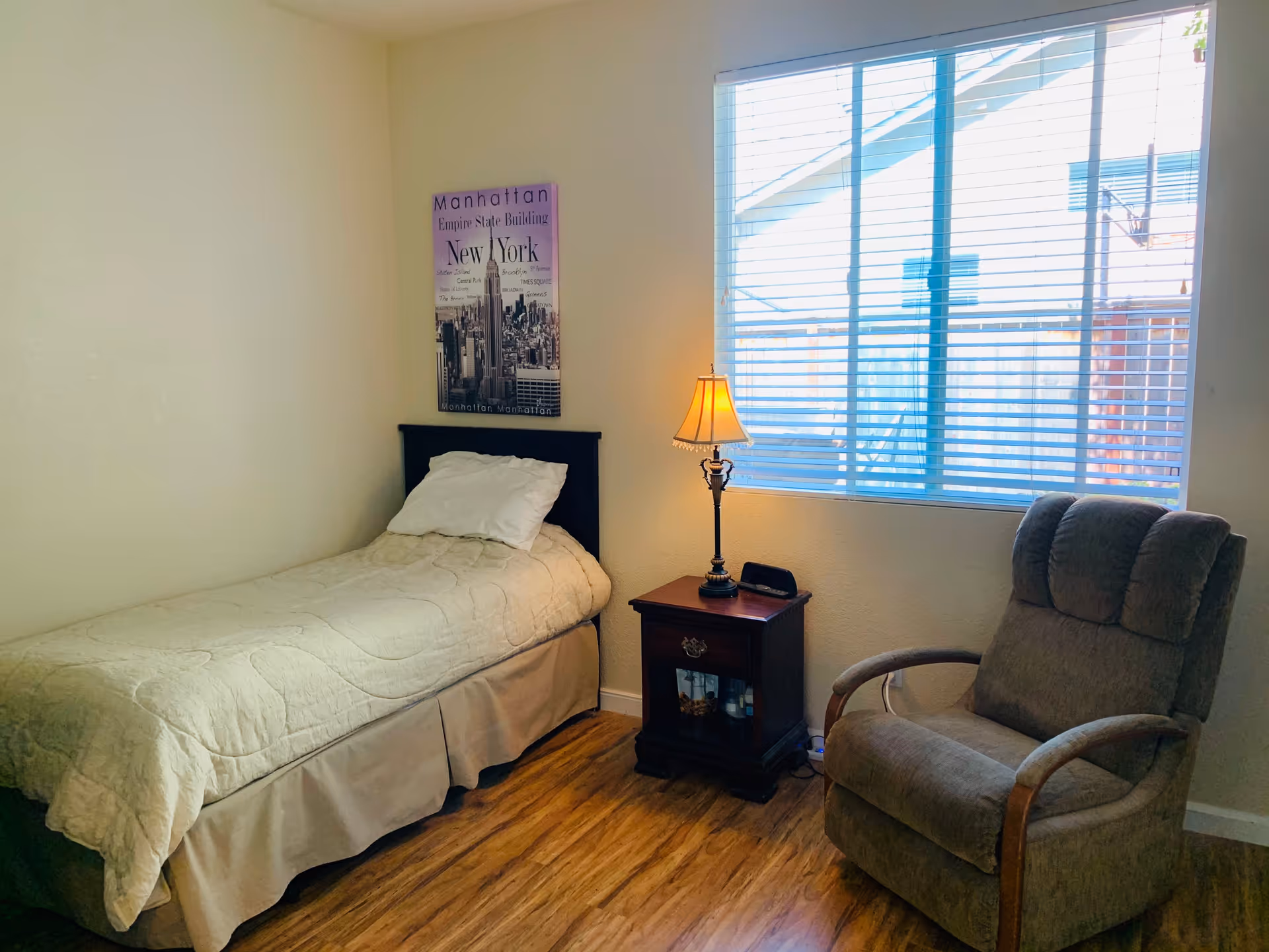 A small bedroom with a single bed covered with a beige quilt and a white pillow. Next to the bed is a wooden nightstand with a decorative lamp and a small clock. A large window with white blinds lets in natural light. A comfortable upholstered armchair is positioned near the window. A framed picture of Manhattan, New York hangs on the wall above the bed.