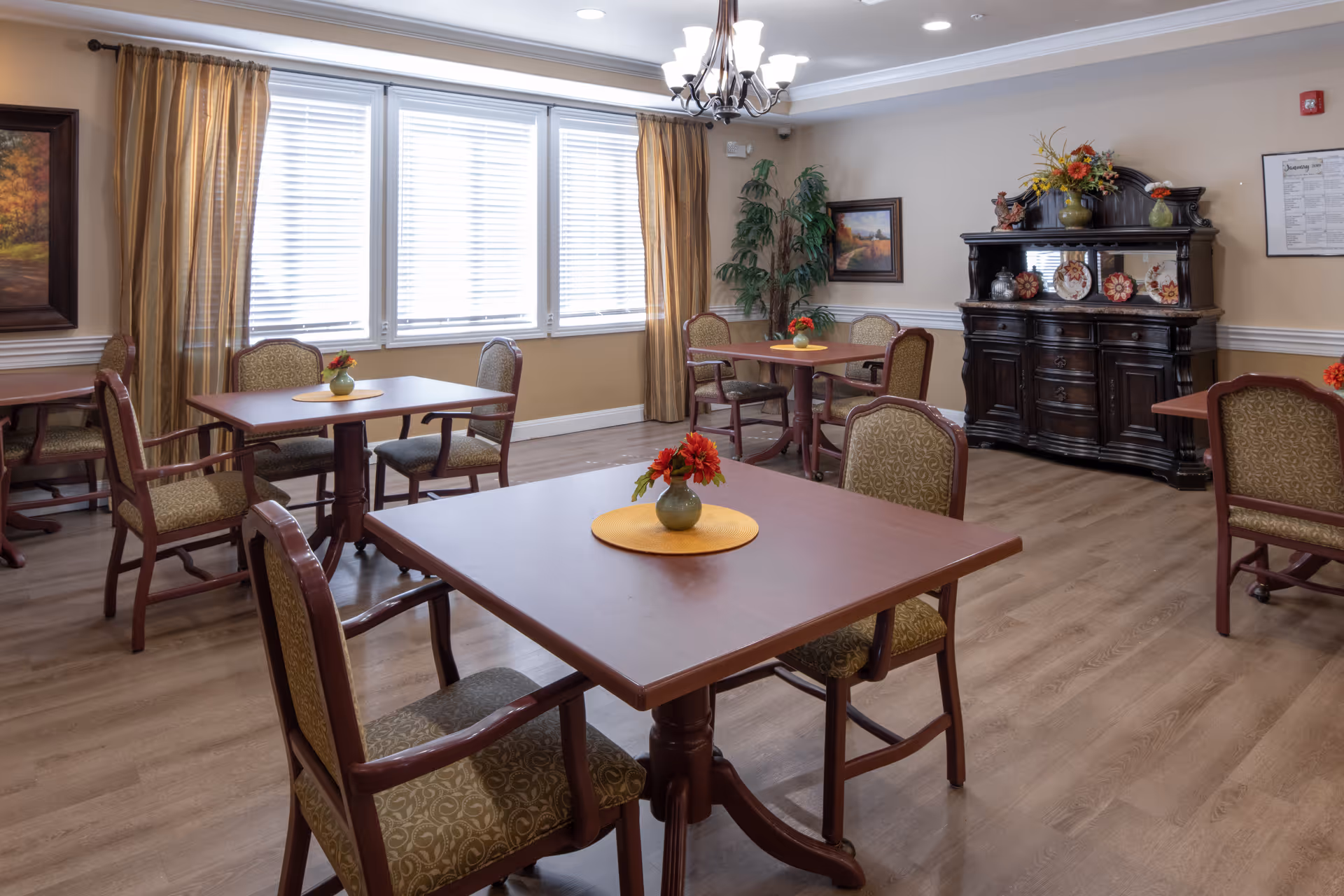 A bright dining room with multiple wooden tables and upholstered chairs, small vases of flowers, large windows with blinds, and a decorative sideboard.