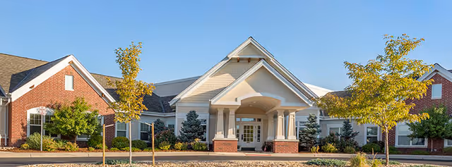 Front exterior view of Brookdale Highlands Ranch facility with a covered entrance, brick and siding facade, landscaped trees and shrubs, and clear blue sky.