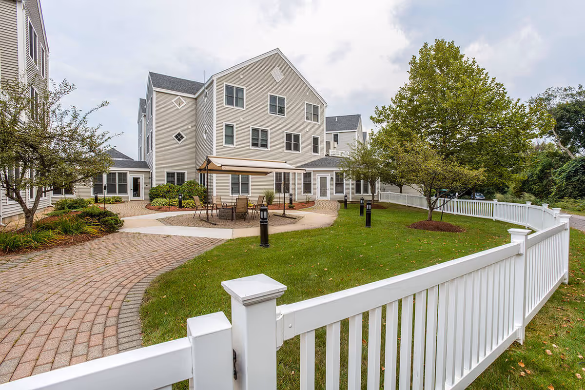 Outdoor courtyard area of a senior living facility with a white fence surrounding a green lawn, several trees, a paved walkway, and a shaded seating area with chairs and a table. The building in the background is beige with multiple windows and three stories tall.