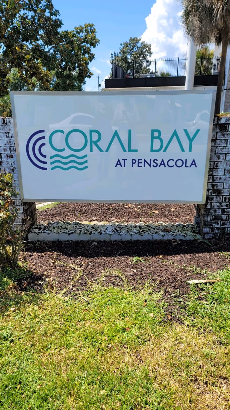 Outdoor sign reading "Coral Bay at Pensacola" mounted between low brick pillars on a grassy lawn.