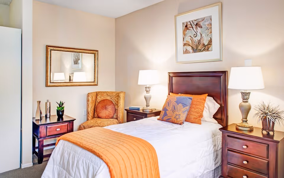 A cozy bedroom with a single bed featuring white and orange bedding, two wooden nightstands with lamps, a decorative armchair, a framed mirror on the wall, and a framed artwork above the bed.