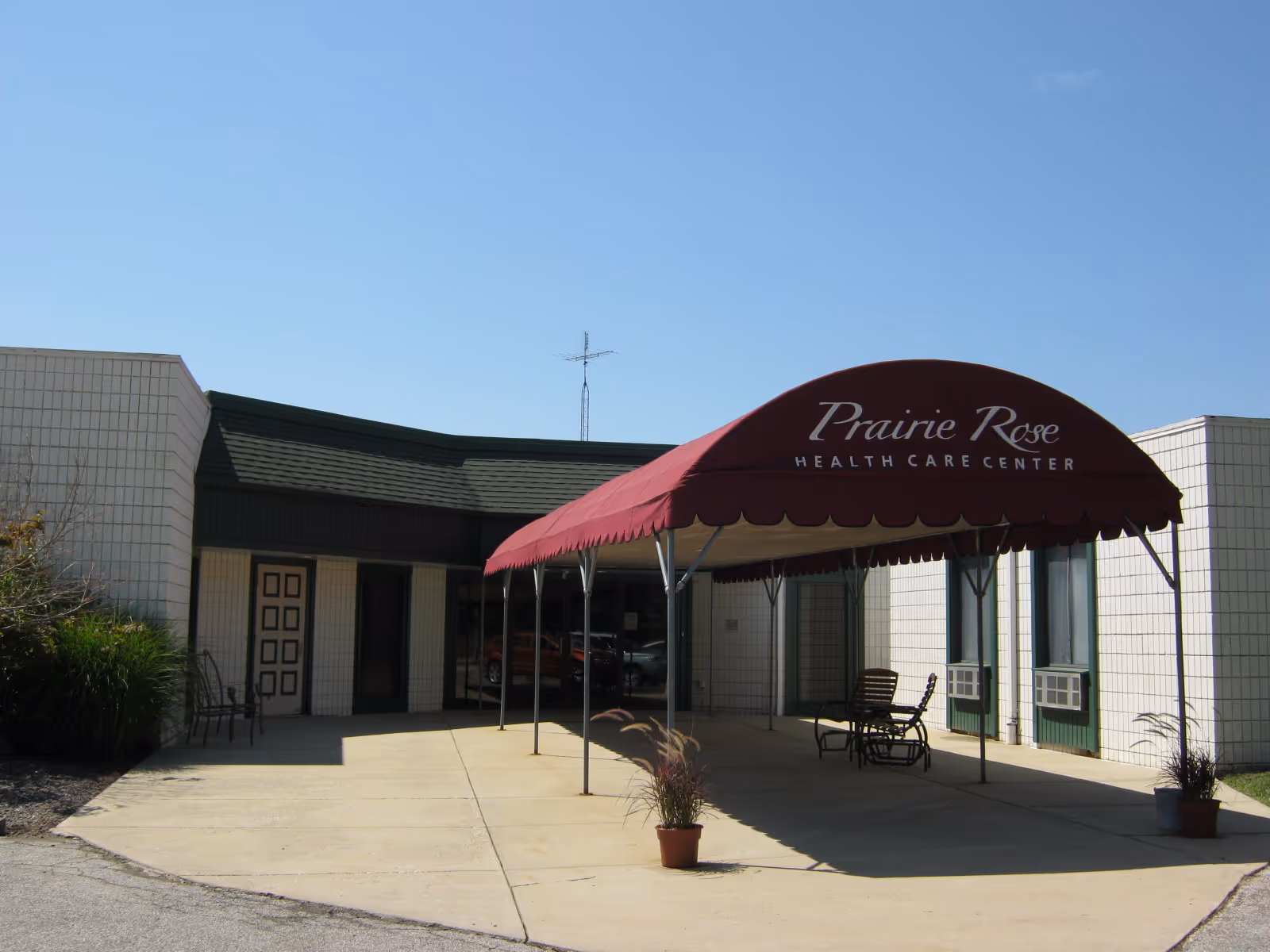 Entrance of Prairie Rose Health Care Center with a maroon canopy covering the driveway. The building is light-colored with a dark green roof trim. There are a few chairs and potted plants near the entrance under the canopy.