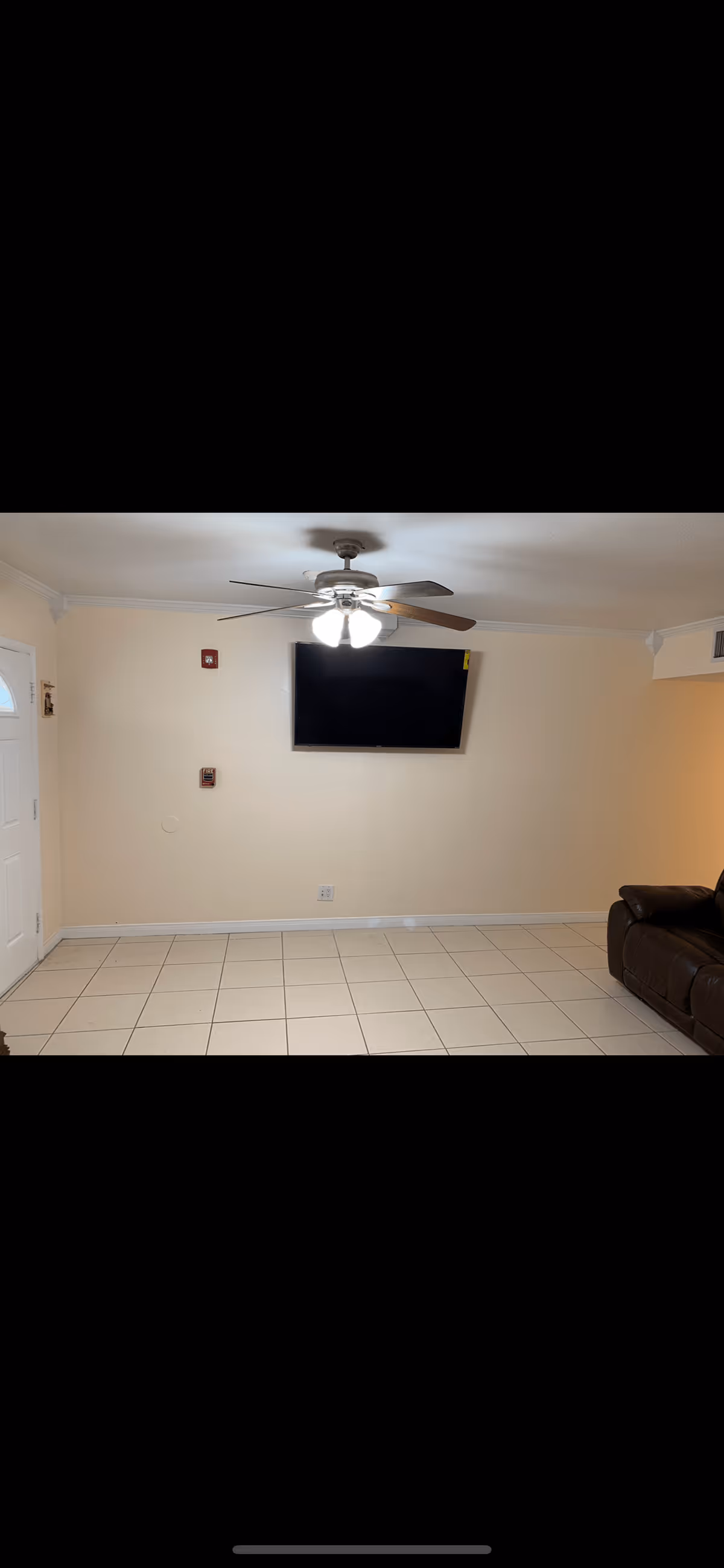 Simple living room with tiled floor, ceiling fan, wall-mounted TV, and a partial brown sofa.