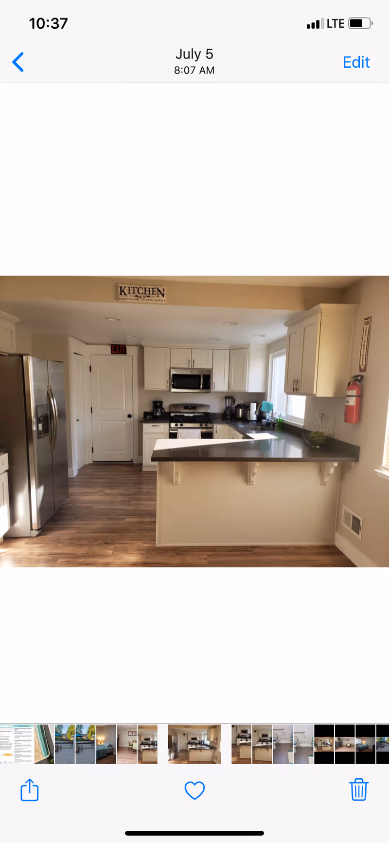 A modern kitchen with white cabinets, stainless steel refrigerator, microwave, and stove. There is a countertop with an overhang for seating, a window letting in natural light, and a fire extinguisher mounted on the wall. The floor is wood, and there is a sign above the kitchen area that reads 'KITCHEN'.