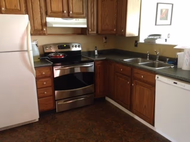 A kitchen with wooden cabinets, a white refrigerator, a stainless steel electric stove with a red pan on it, a double sink, and a white dishwasher. The countertops are dark, and there is a framed picture on the wall above the sink area.