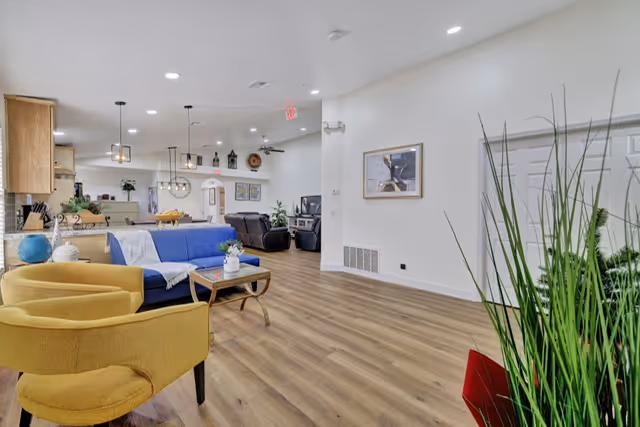 A spacious and modern living area with light wood flooring, white walls, and recessed lighting. The room features a blue sofa, two yellow armchairs, a wooden coffee table with a small plant, and a kitchen area in the background with hanging pendant lights. There are decorative plants and framed artwork on the walls.