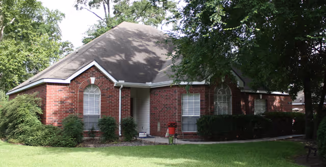 Brick cottage-style building with a front entrance, shrubs, and large trees.