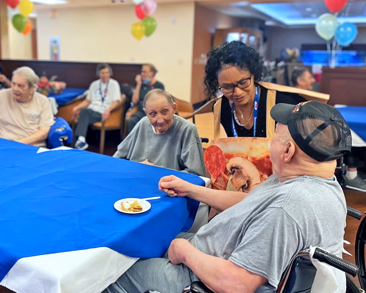 A group of elderly people seated around a table covered with a blue tablecloth in a communal dining or activity room. A woman wearing glasses and a pizza-themed apron is engaging in conversation with a man in a wheelchair wearing a black cap. Balloons in various colors are visible in the background, adding a festive atmosphere.