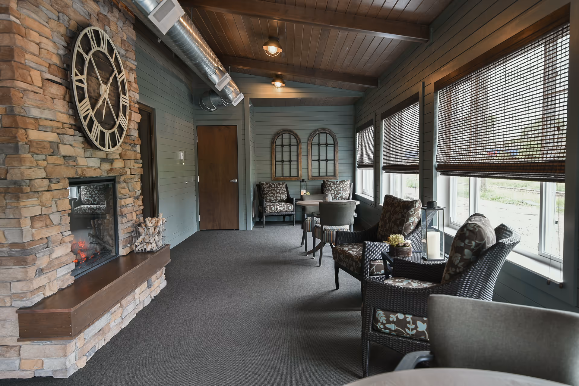 Sunlit lounge with a stone fireplace, wicker armchairs, patterned cushions and large windows with bamboo blinds.