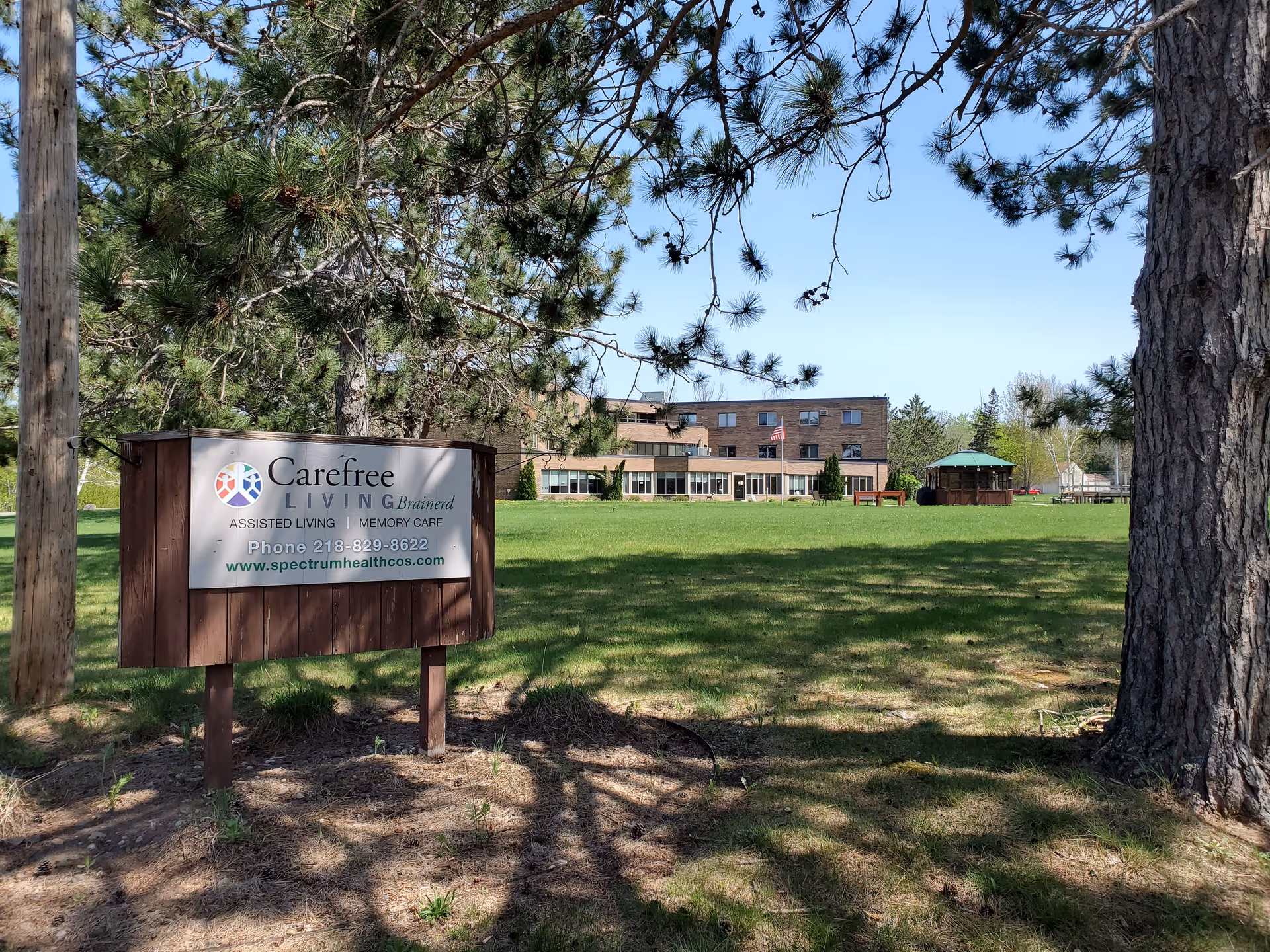 Outdoor view of Brainerd Carefree Living facility with a large green lawn, trees, and a wooden sign in the foreground displaying the facility's name, services, phone number, and website. The building is visible in the background with an American flag and a gazebo on the lawn.