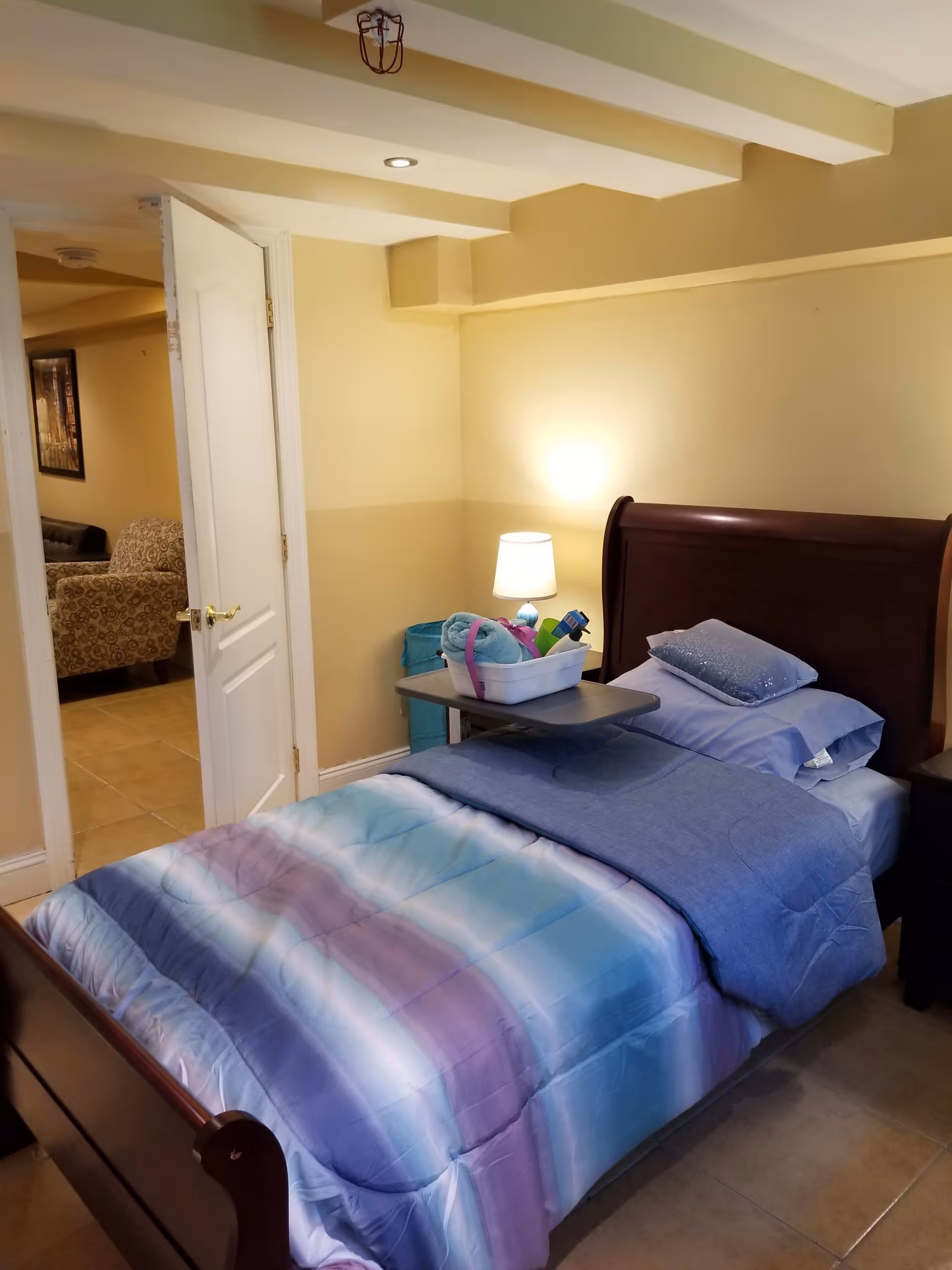 A bedroom in an assisted living facility featuring a single bed with a wooden headboard and footboard. The bed is made with a blue and purple striped comforter, blue sheets, and a pillow. A small bedside table with a lamp and a tray holding towels and toiletries is next to the bed. The room has beige walls and tile flooring. An open door reveals a glimpse of a living area with patterned armchairs and a framed picture on the wall.