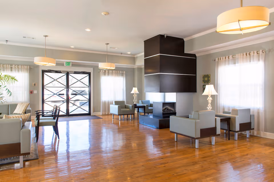 A spacious and well-lit common area with wooden flooring, modern light fixtures, and large windows with sheer curtains. The room features multiple seating arrangements with light gray armchairs and small tables, centered around a contemporary black fireplace. The entrance door with glass panels is visible in the background.