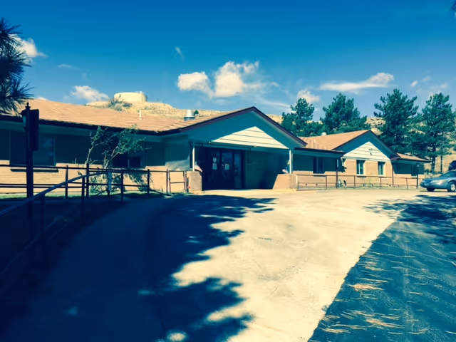 Exterior view of a single-story building with a sloped roof, surrounded by trees and a clear blue sky. The building has a covered entrance with double glass doors and a paved driveway leading up to it.