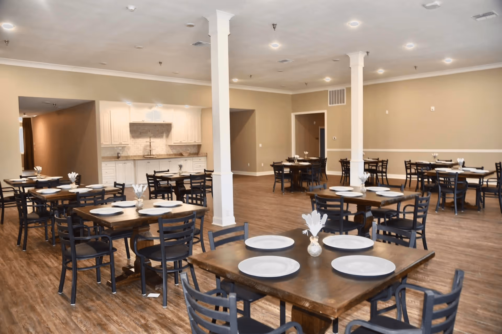 A spacious dining room with multiple wooden tables and black chairs arranged neatly. Each table is set with white plates and small decorative centerpieces. The room has beige walls, wooden flooring, white pillars, and recessed ceiling lights. In the background, there is a white kitchen area with cabinets and a countertop.