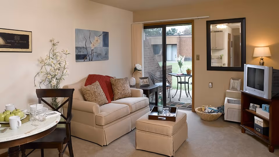 Cozy living room with a loveseat, ottoman, small dining table, TV cabinet, and sliding glass door opening to a patio.