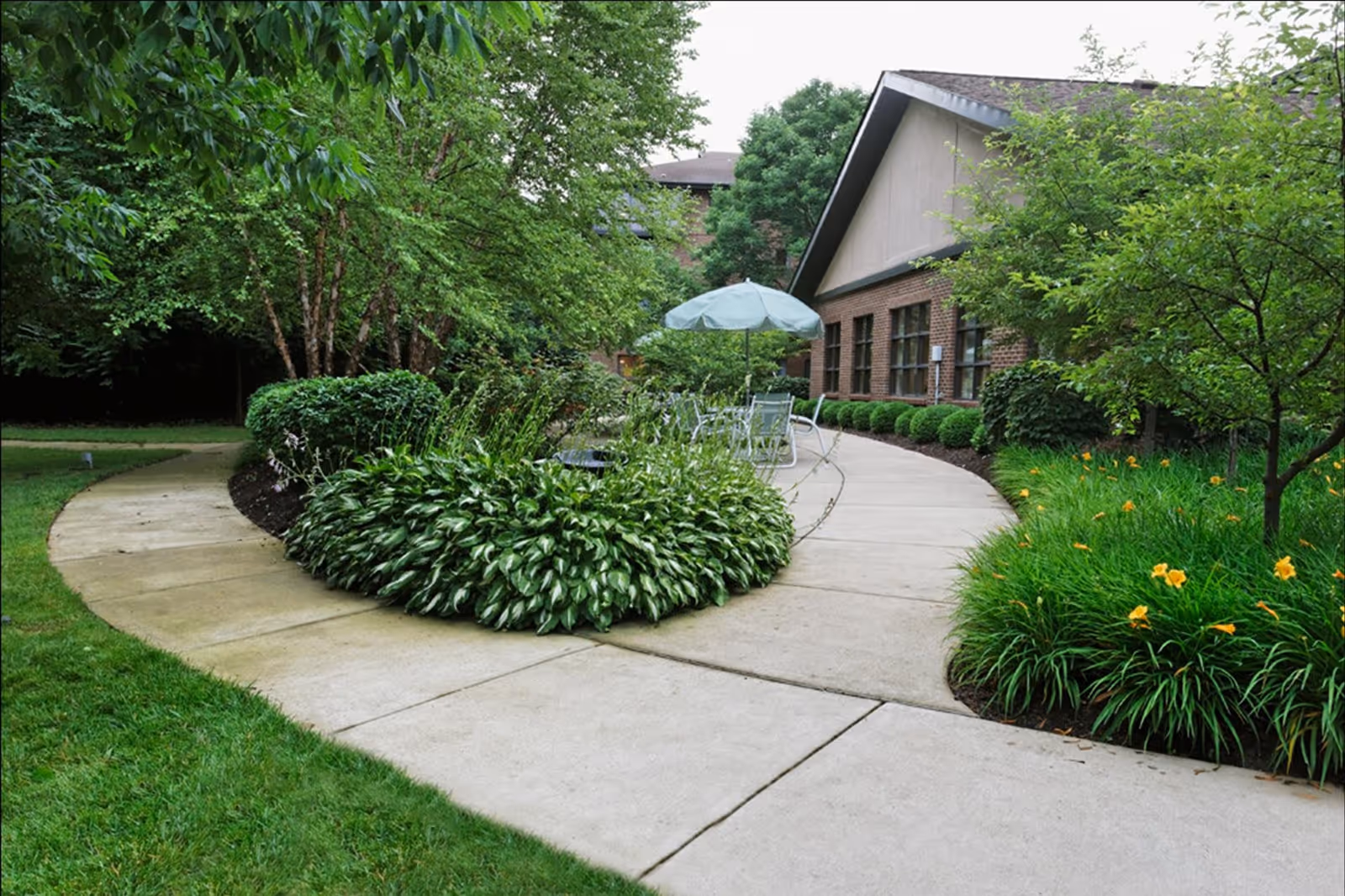 Curved concrete walkway surrounded by lush green plants and trees leading to a brick building with large windows. There is a patio area with a table, chairs, and a blue umbrella.