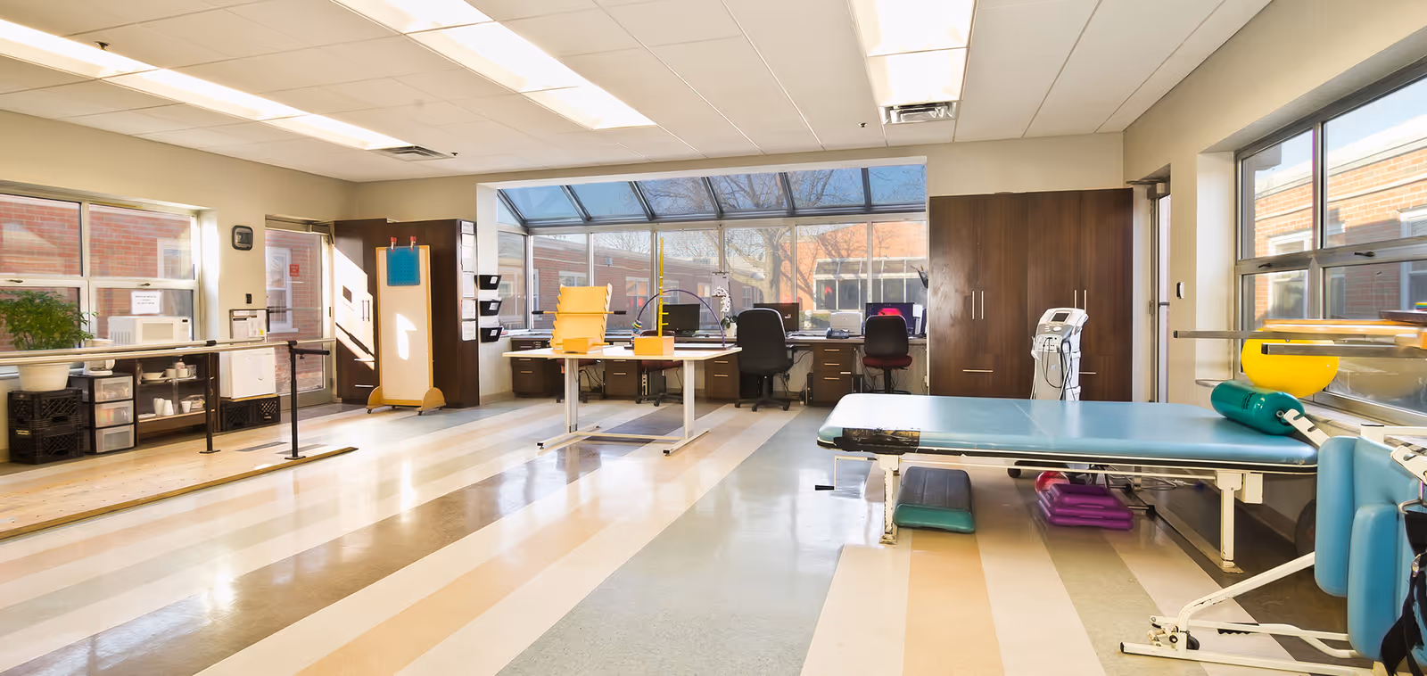 A spacious therapy or rehabilitation room with large windows allowing natural light. The room contains a therapy table with various exercise equipment, two office chairs at desks with computers, storage cabinets, and a whiteboard. The floor has wide striped patterns in beige, brown, and light blue tones.
