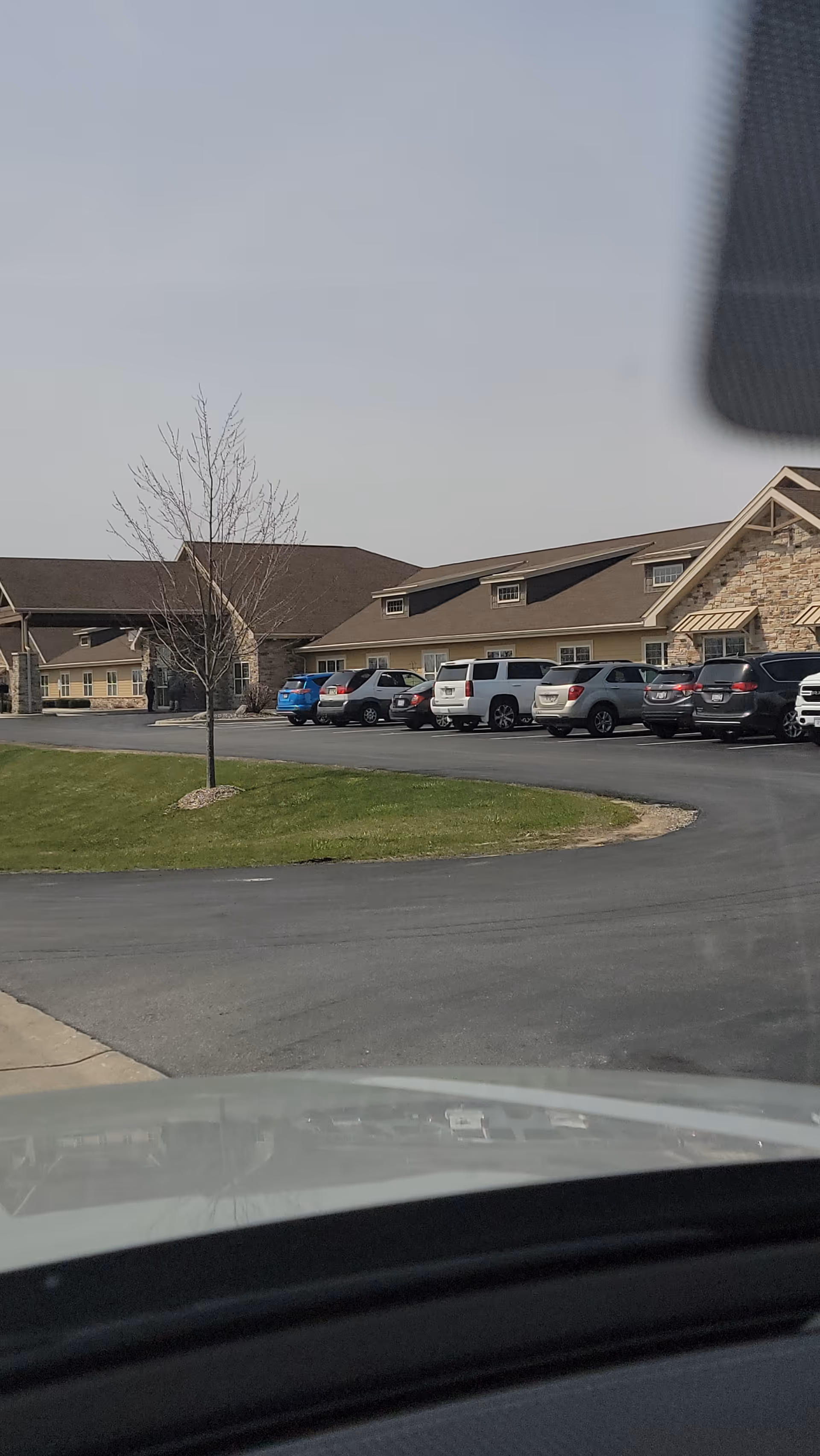 View of the exterior of a senior care facility building with a parking lot in front. Several cars are parked along the building, which has a stone and beige facade with a sloped roof. A small tree is planted on a grassy area near the driveway.