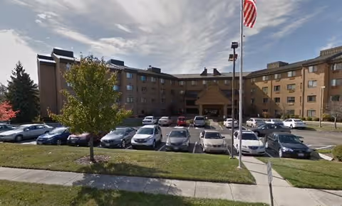 Front exterior of a multi-story brick skilled nursing facility with a parking lot, cars, and an American flag at the entrance.